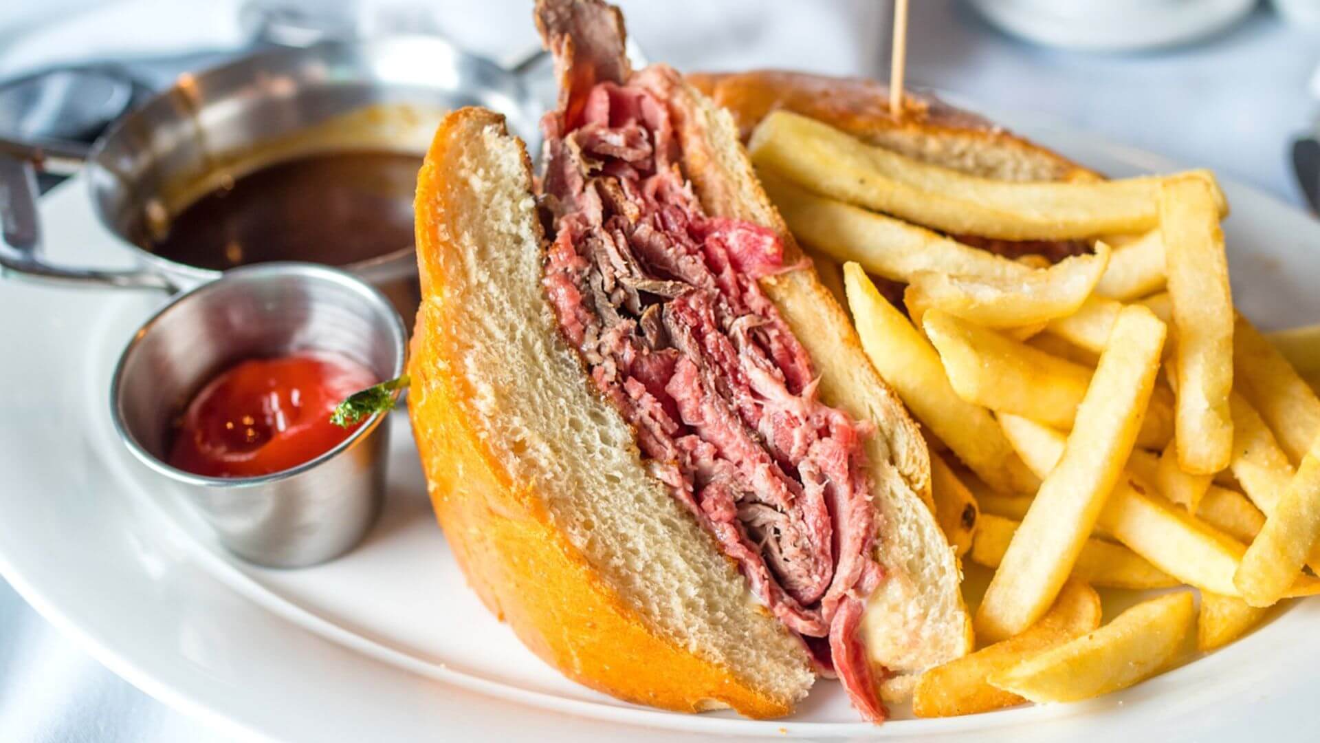 A roast beef sandwich cut in half to reveal layers of thinly sliced meat, served on a white plate with a side of french fries, a small cup of ketchup, and a bowl of dark dipping sauce.