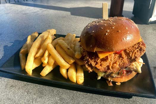 A crispy fried chicken burger with melted cheese and lettuce on a sesame brioche bun, served alongside golden french fries on a black rectangular plate.